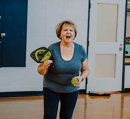 An older woman yells across a gymnasium while holding a pickleball racket and ball.