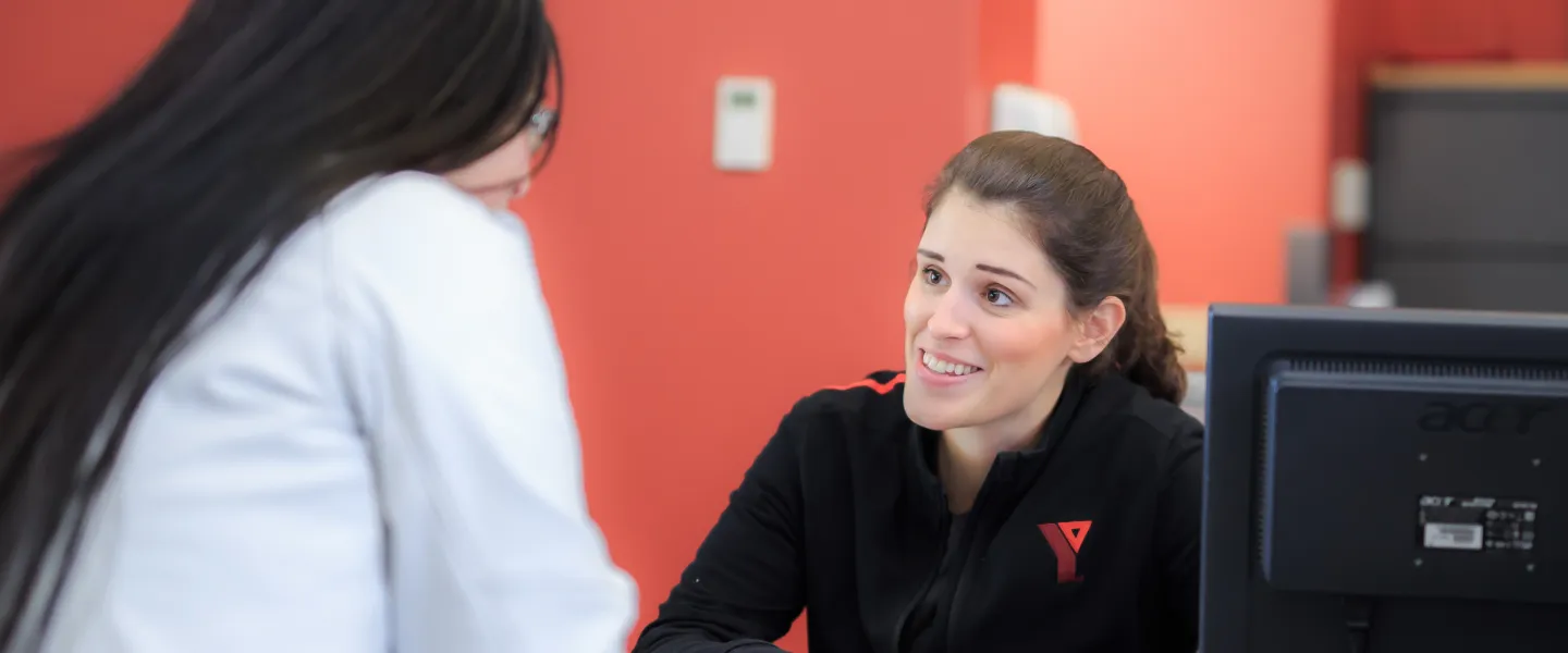 A YMCA staff member sits behind a customer service desk, talking to a guest about programs and services.
