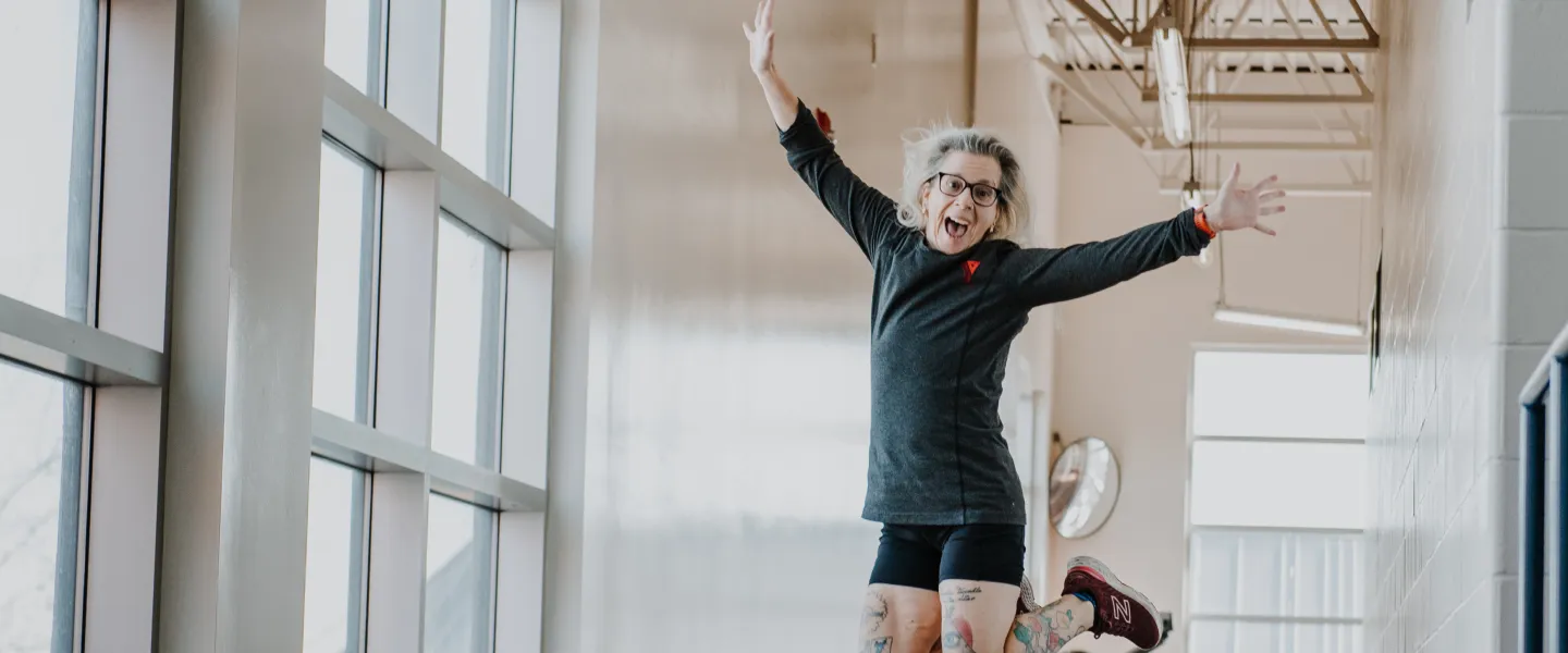 A young woman wearing a grey long-sleved shirt seen with a YMCA logo jumps in the air while on an indoor walking track.