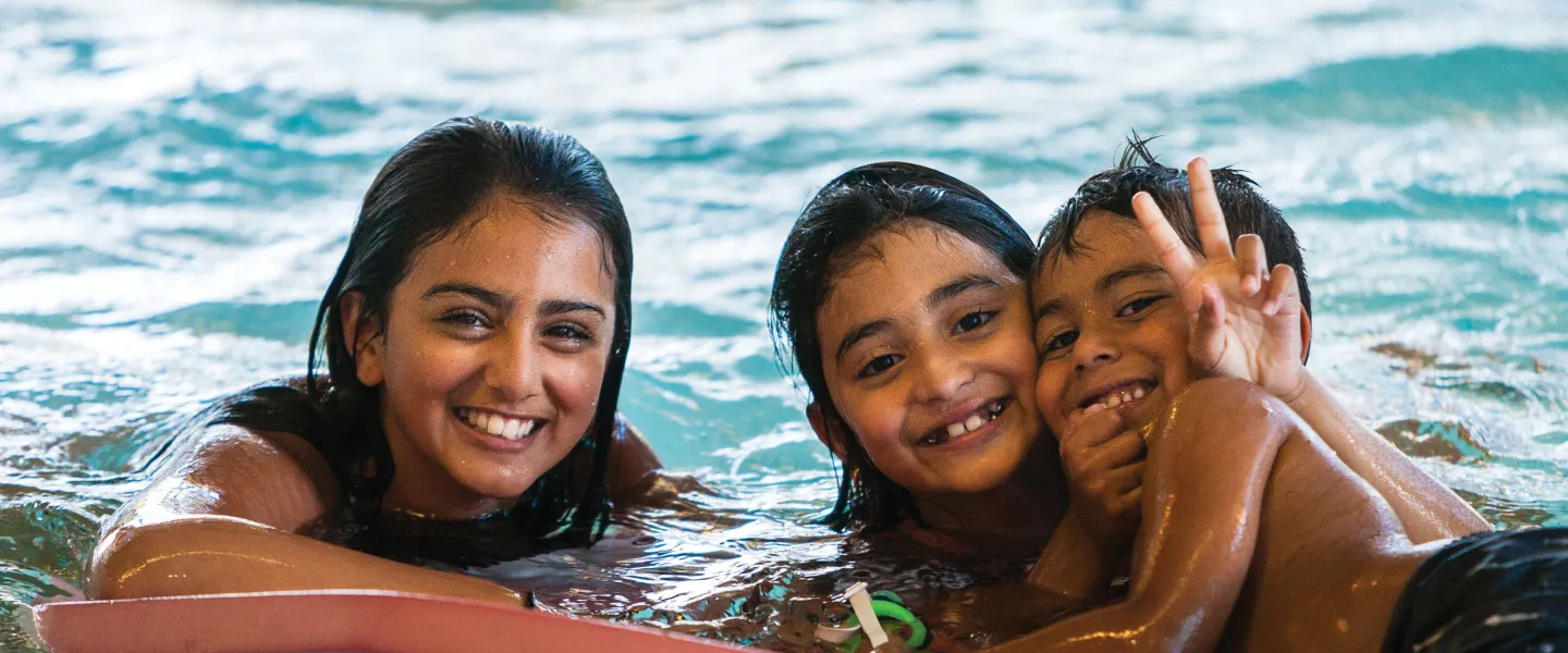 Three kids in a pool hold onto a toy and pose for a photo.