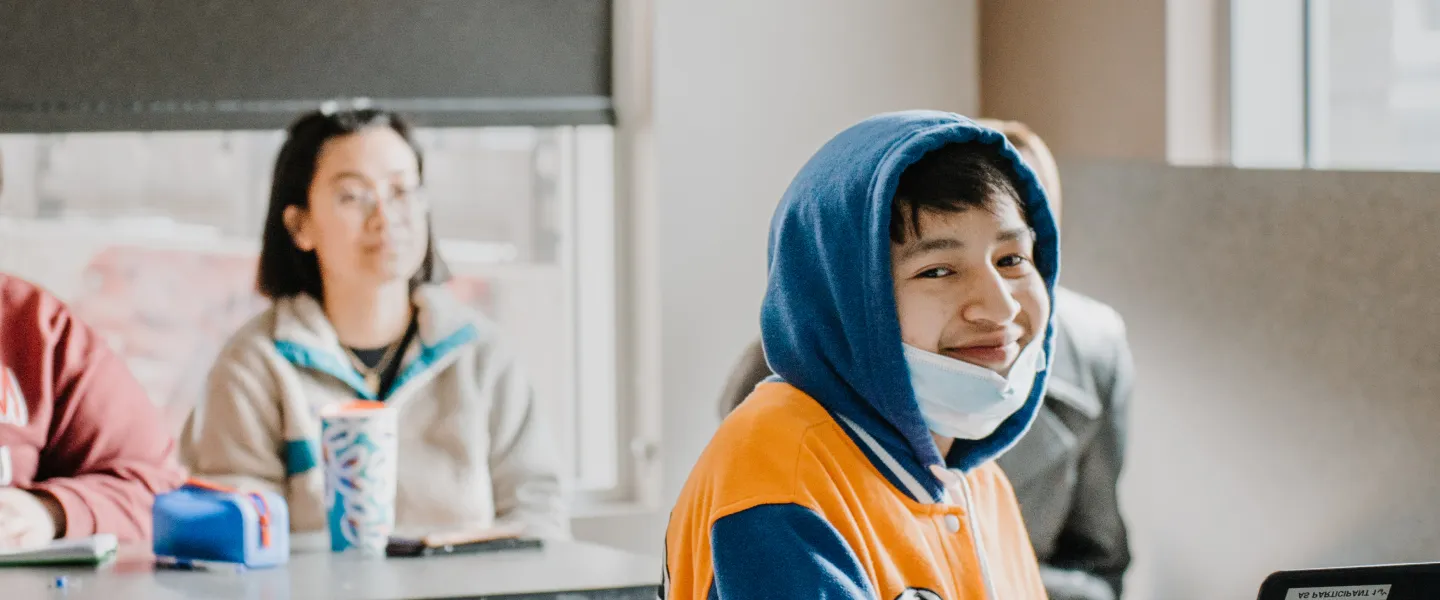 A teenage boy wears a hoodie with the hood up, sits at a table in a classroom with a laptop open.