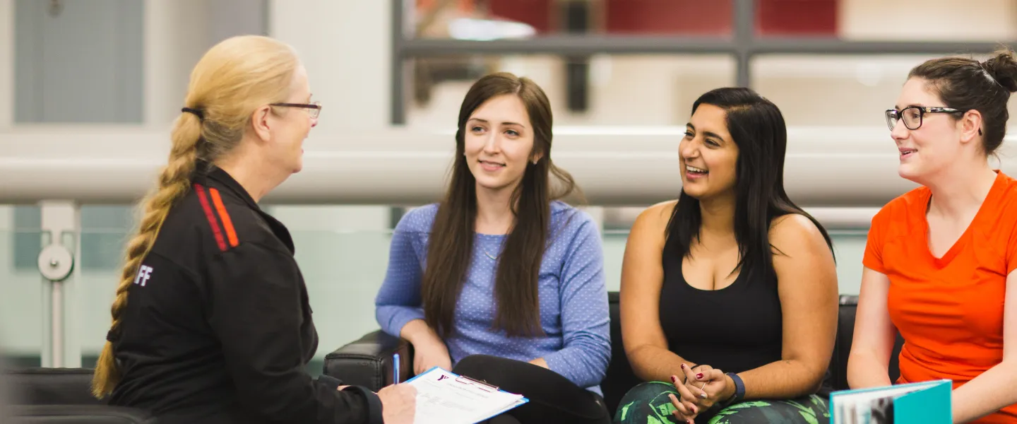 A YMCA staff member holds a clipboard while she talks to three young women on couches at a YMCA.