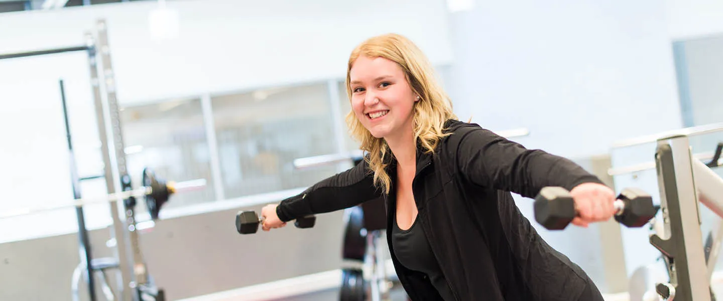 A smiling young lady working out at gym holding dumbbells..