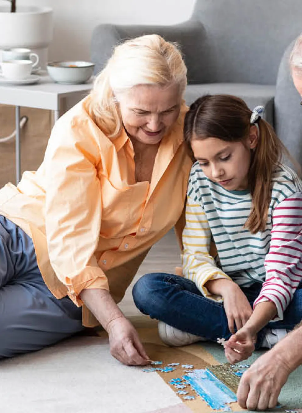 A grandmother, granddaughter and grandfather put a puzzle together on their livingroom floor.
