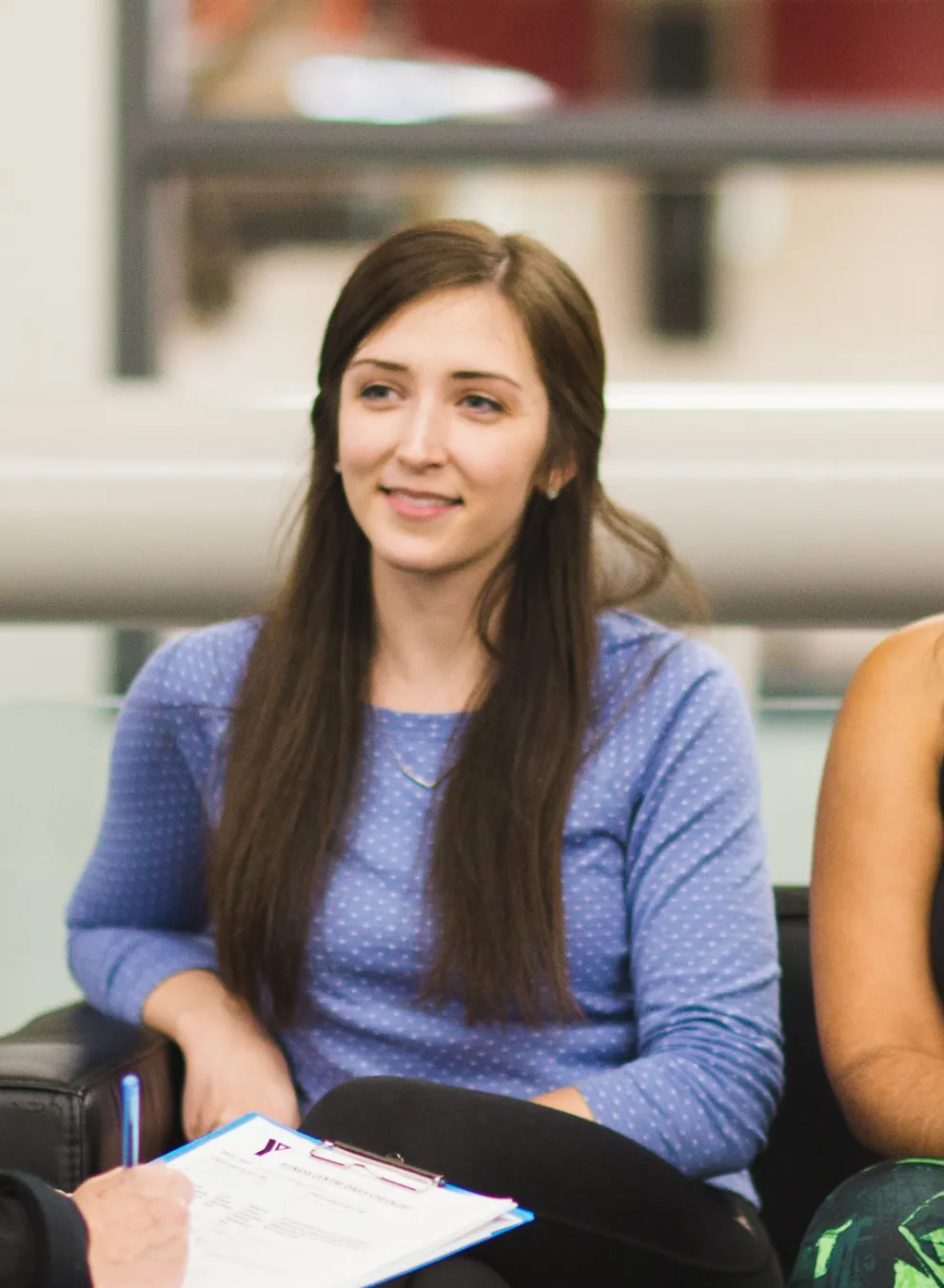 A YMCA staff member holds a clipboard while she talks to three young women on couches at a YMCA.