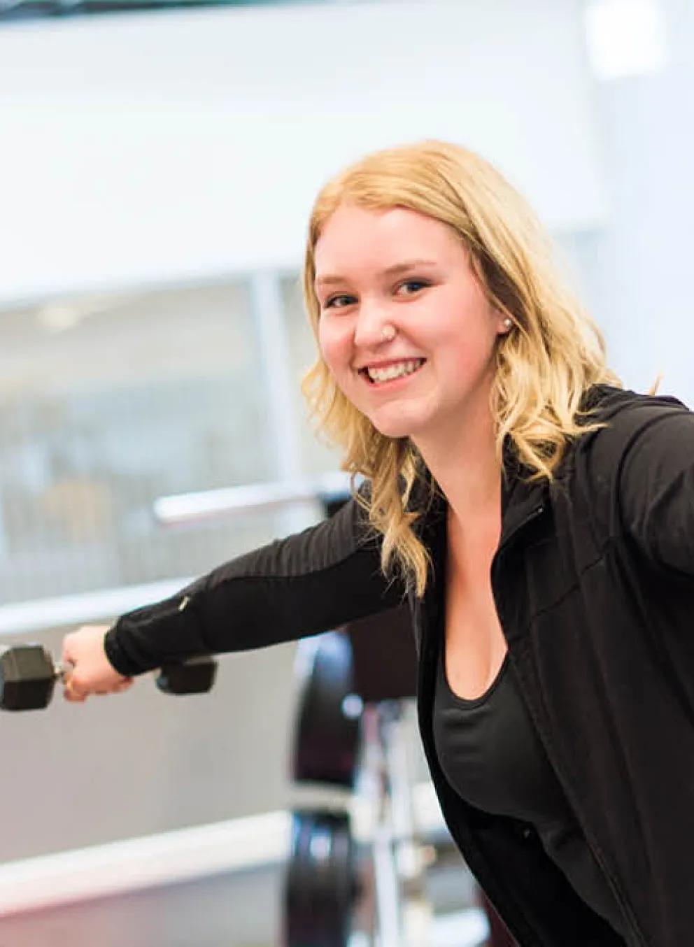 A smiling young lady working out at gym holding dumbbells..