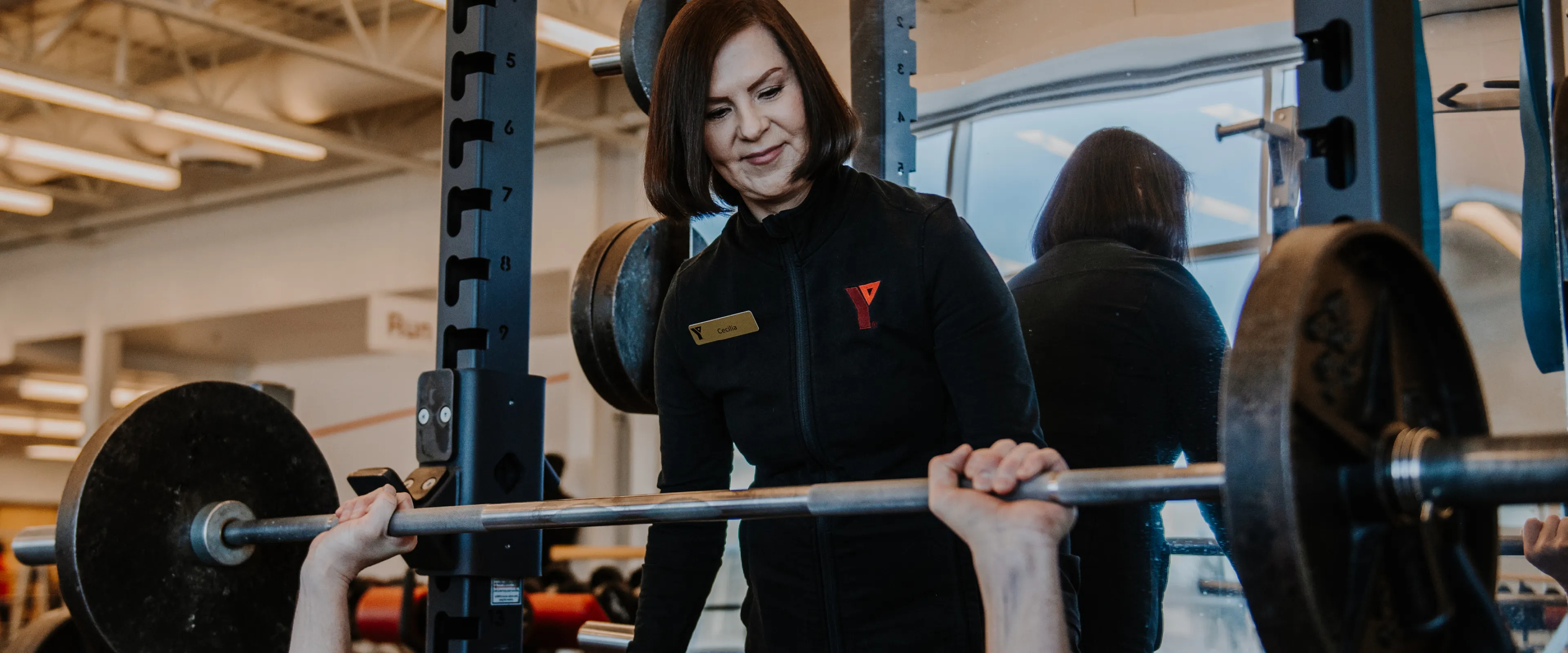 A female personal trainer stands behind a male member as he uses a barbell bench press.