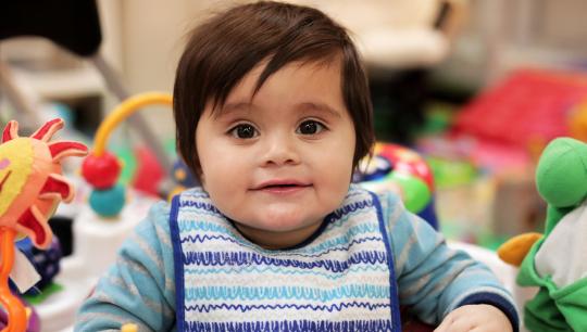 A baby wears a striped shirt and artisitic bib while sitting in an exersaucer.