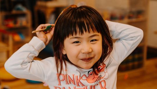 A young girl wears a long-sleved Pink Panther shirt while hiding a toy behind her head.