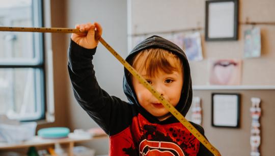 A young boy wears his hood up on his head while playing with a measuring tape.