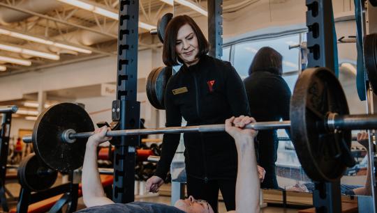 A female personal trainer stands behind a male member as he uses a barbell bench press.