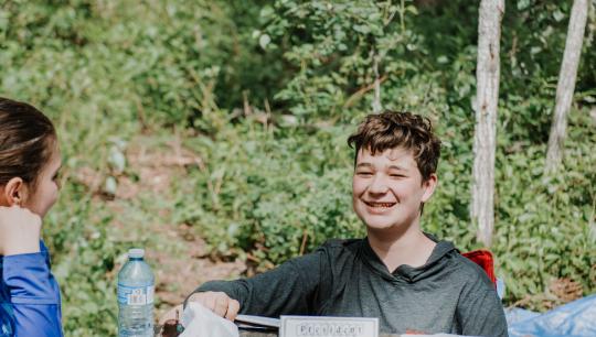 A group of teens play a card game on a wooden picknic table while camping.