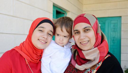 A teenage girl, young boy and their mother stand on their front porch, posing for a photo.