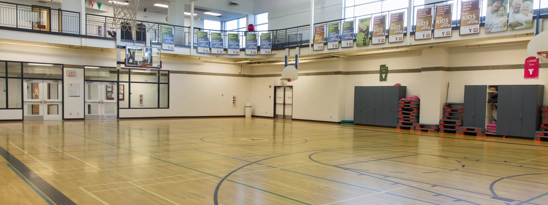 A large gymnasium featuring wood flooring and plently of light shining through second-story windows.