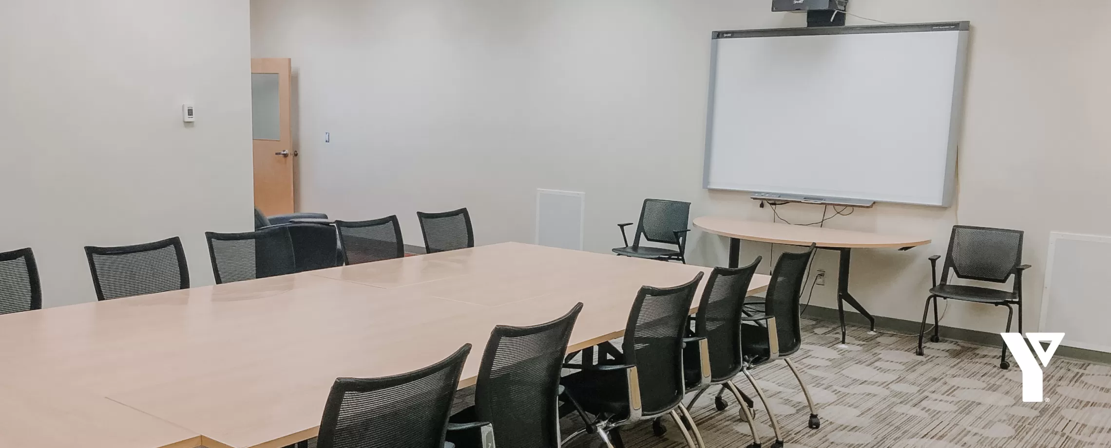 A brightly lit room featuring a SMART board attached to a wall, with a large table with numerous chairs around it in the middle.