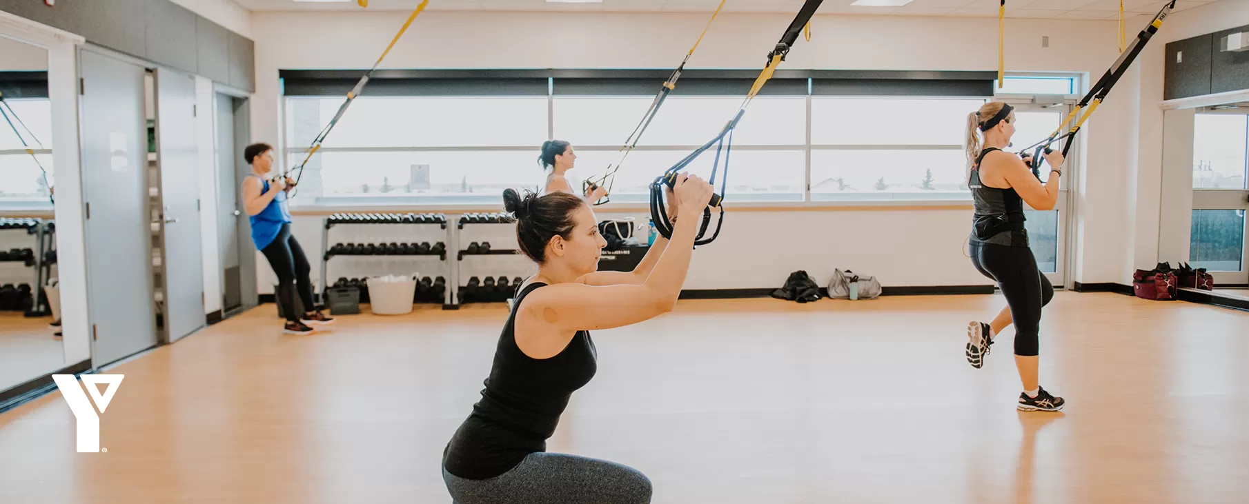 A small group fitness class uses TRX equiptment in a sun-filled studio space.