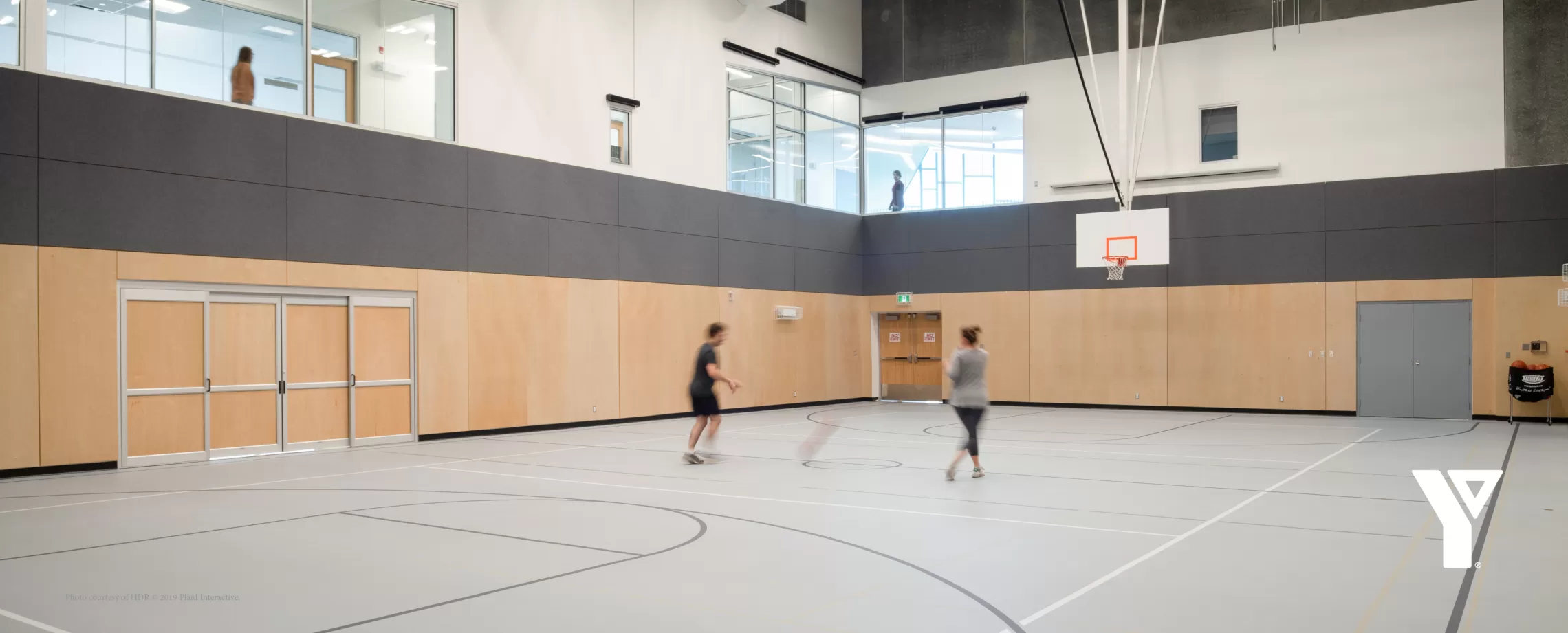 Two people play basketball in a large, sun-filled gymnasium.