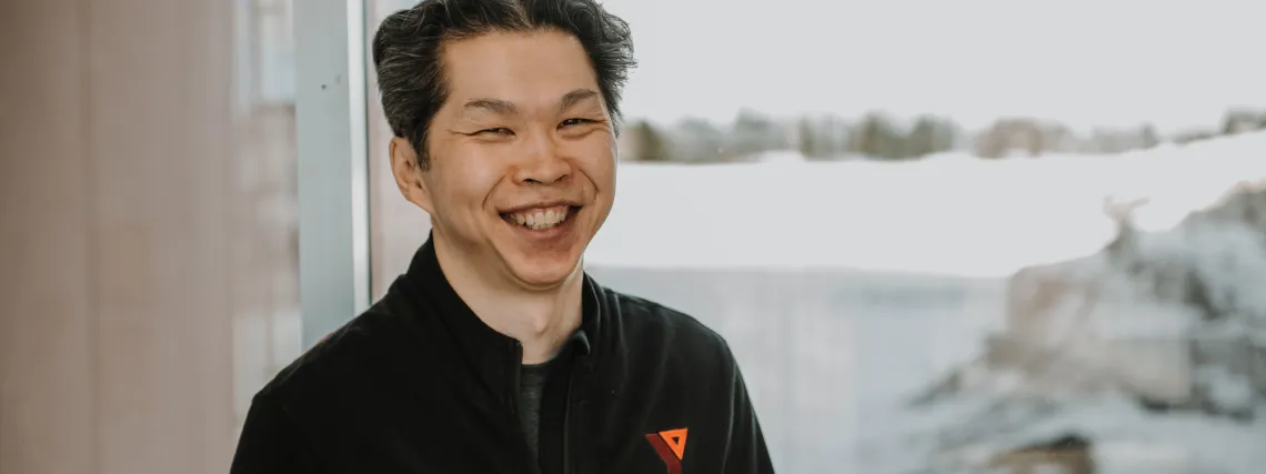 An Asian man wears a black jacket seen with a Y logo and stands infront of a large window.