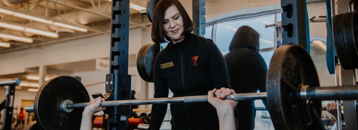 A female personal trainer stands behind a male member as he uses a barbell bench press.