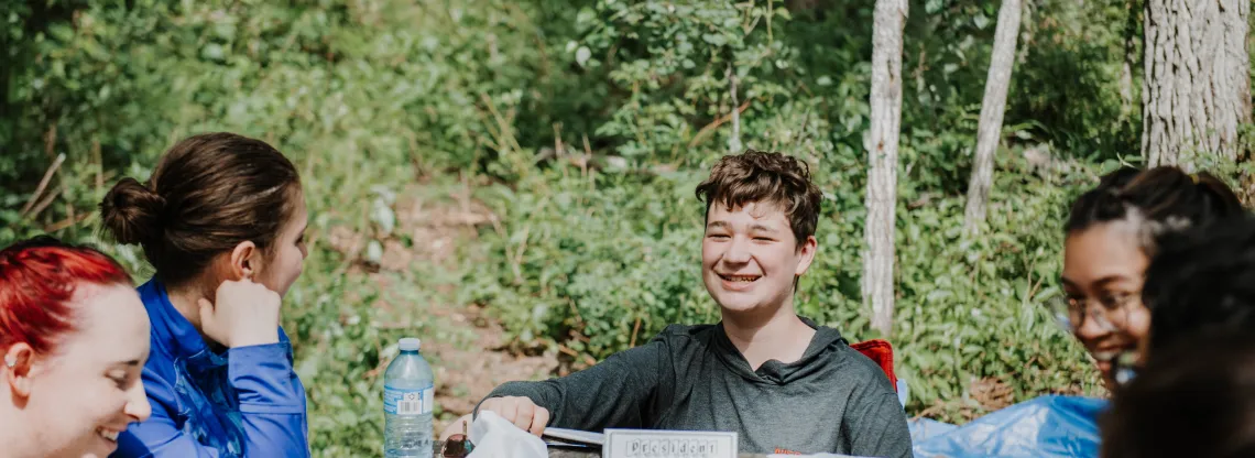 A group of teens play a card game on a wooden picknic table while camping.