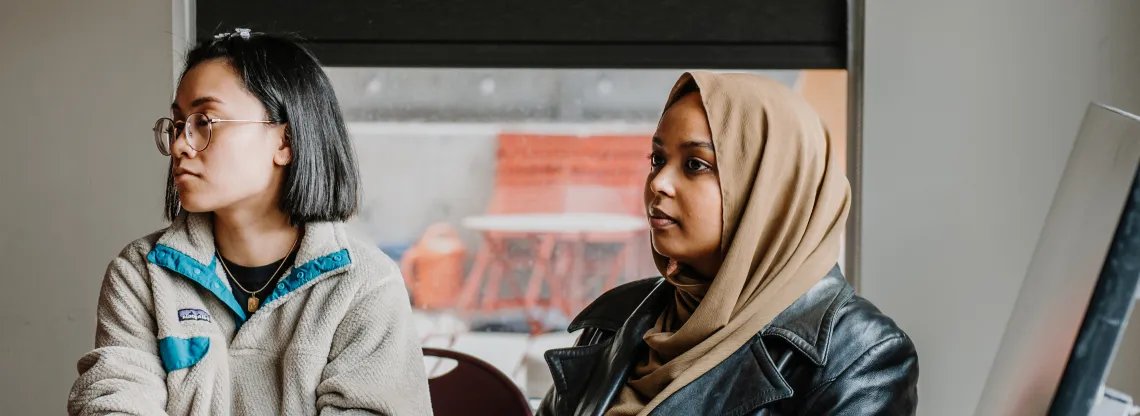 Two young women sit adjasent to each other in a brightly lit room while listening to a workshop.