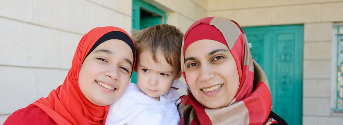 A teenage girl, young boy and their mother stand on their front porch, posing for a photo.